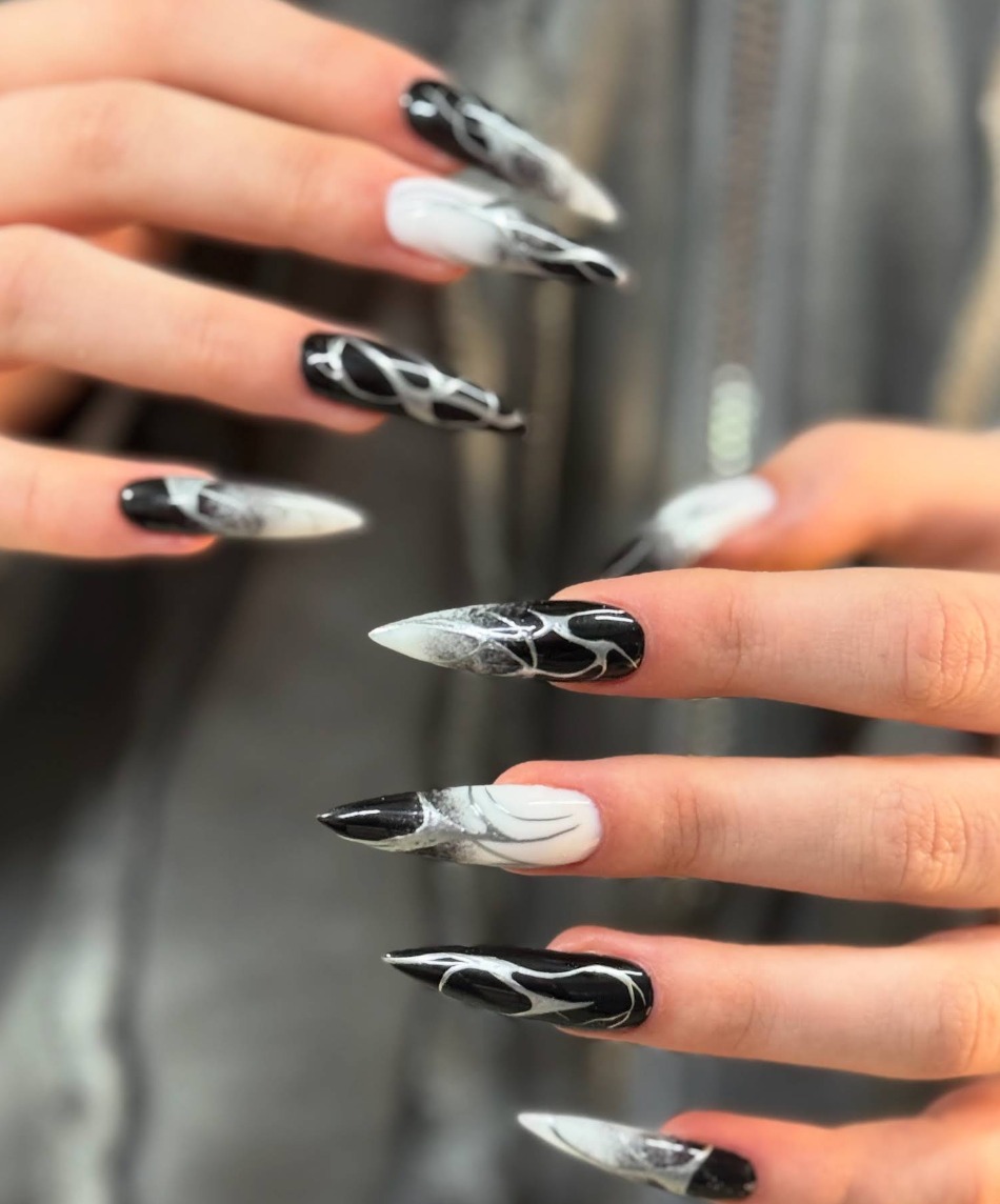 Dłonie z pomalowanymi paznokciami w czarno-białe wzory, niektóre w kształcie płomieni. Część paznokci jest w szpic, a część ma kształt prostokątny.
