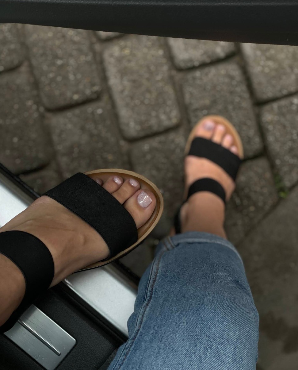 Stopa w sandałach z jasnym, błyszczącym zdobieniem paznokci. Widoczne jeansy i fragment wnętrza samochodu. Kąt z góry.