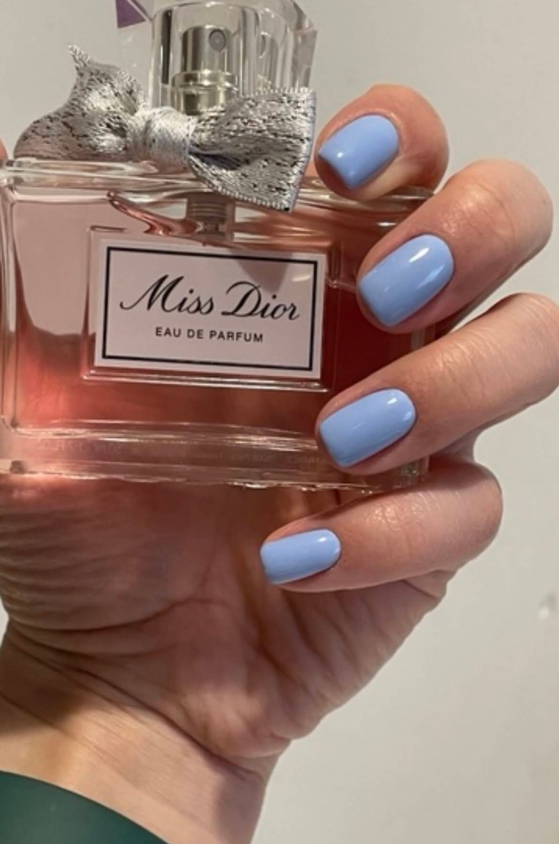 Dłoń z błękitnym manicure hybrydowym na tle flakonu perfum Miss Dior. Delikatny odcień lakieru i elegancki flakon tworzą spójną, estetyczną kompozycję.