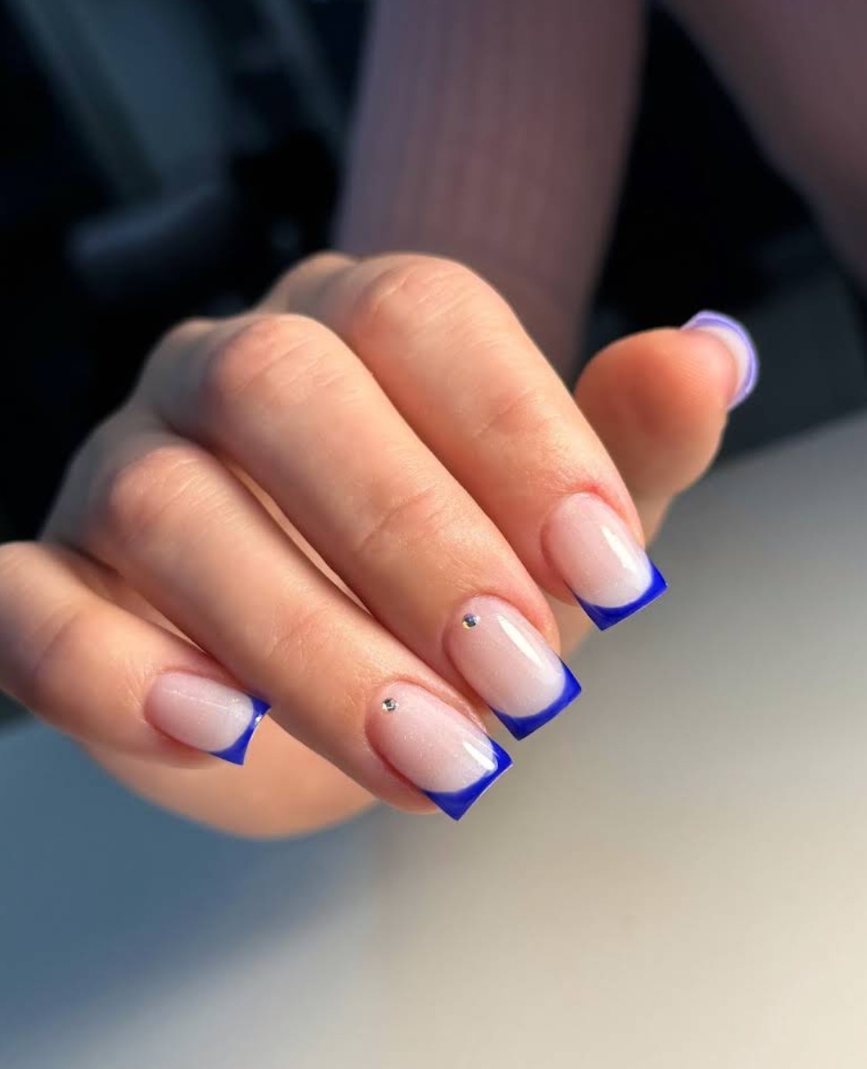 Dłoń z kwadratowymi paznokciami french manicure, z kobaltową końcówką i drobnymi srebrnymi zdobieniami na dwóch palcach. Estetyczne i zadbane dłonie.