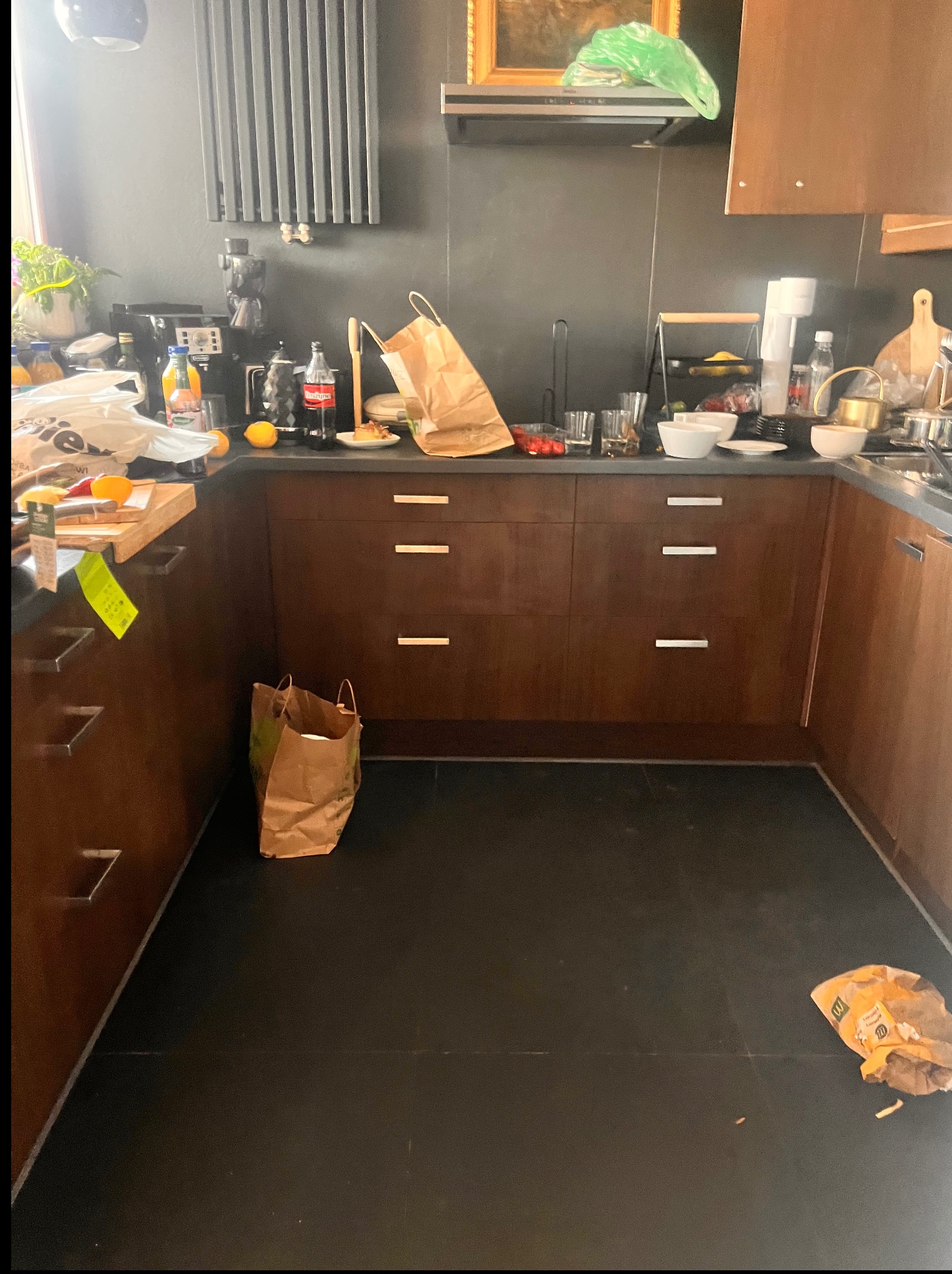 Niefotogeniczna kuchnia z ciemnymi szafkami, na blacie bałagan i torby. Na podłodze śmieci. Widok na wnętrze przed sprzątaniem, ukazujący potrzebę interwencji.