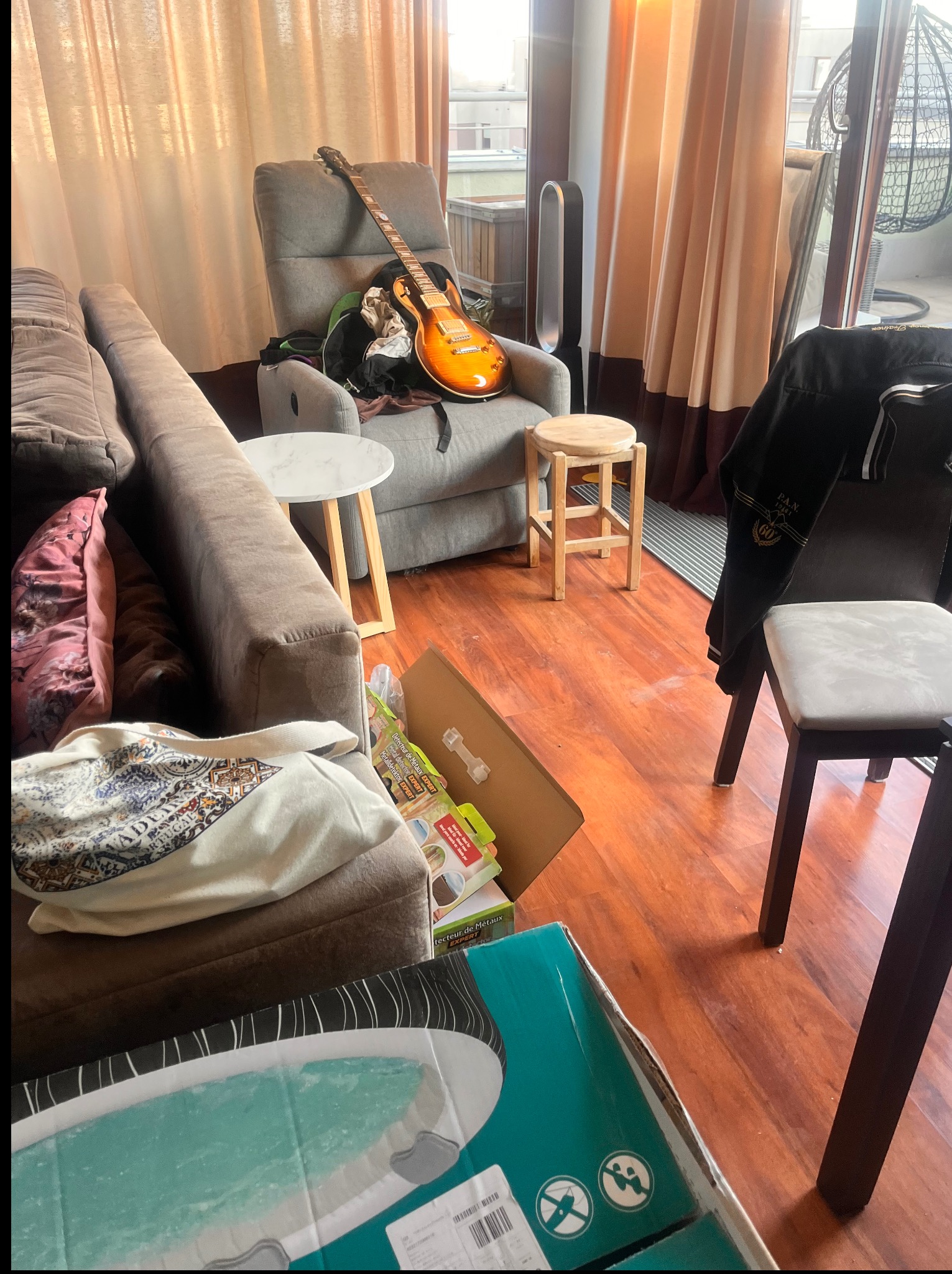 Nieuporządkowany pokój dzienny z gitarą opartą o fotel, rozrzucone przedmioty i kartony na podłodze, sugerujące potrzebę posprzątania.