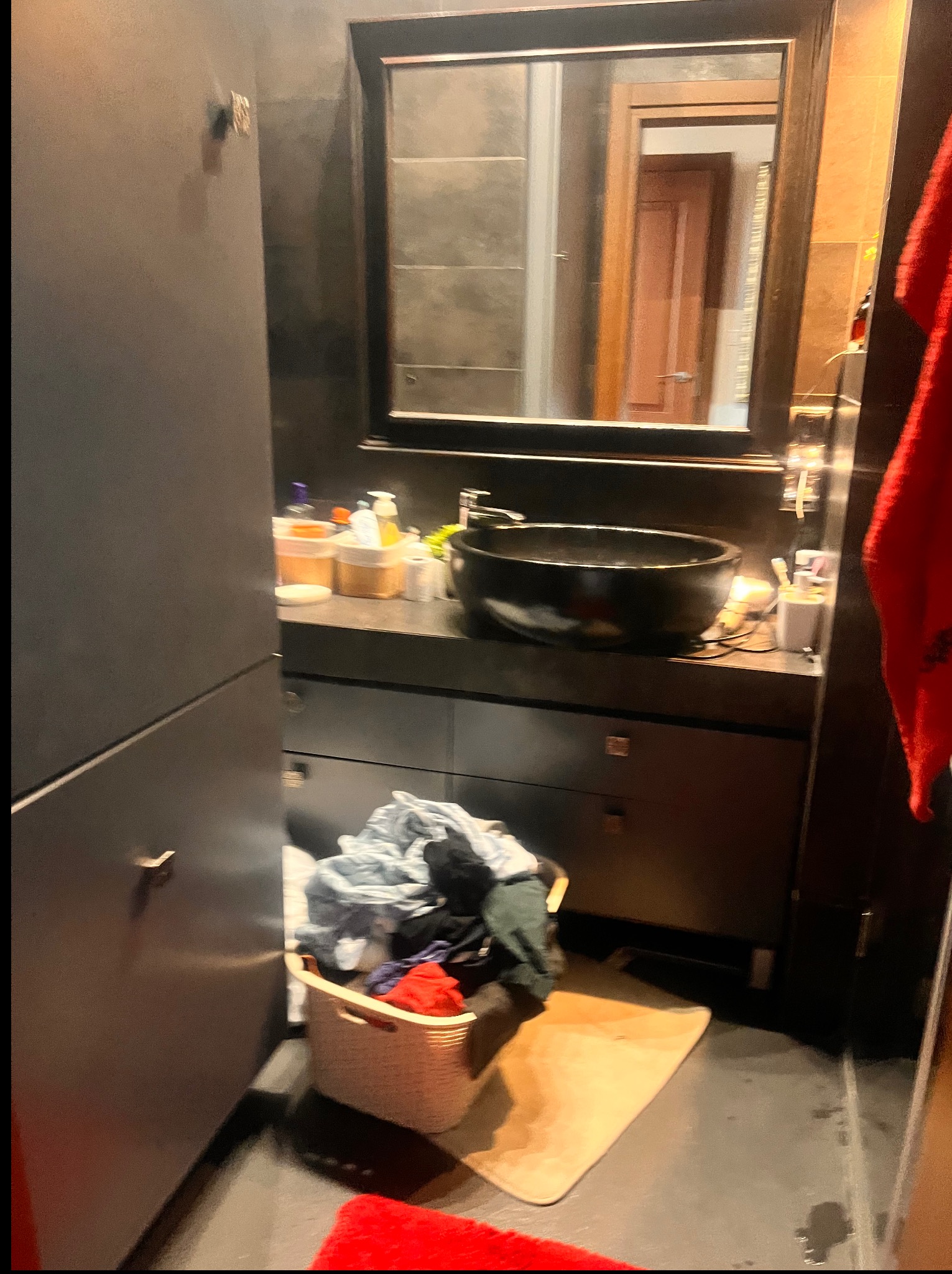 Łazienka z ciemnymi meblami, okrągłą umywalką i koszem z praniem. W lustrze widoczne odbicie innego pomieszczenia. Panuje delikatny nieporządek.