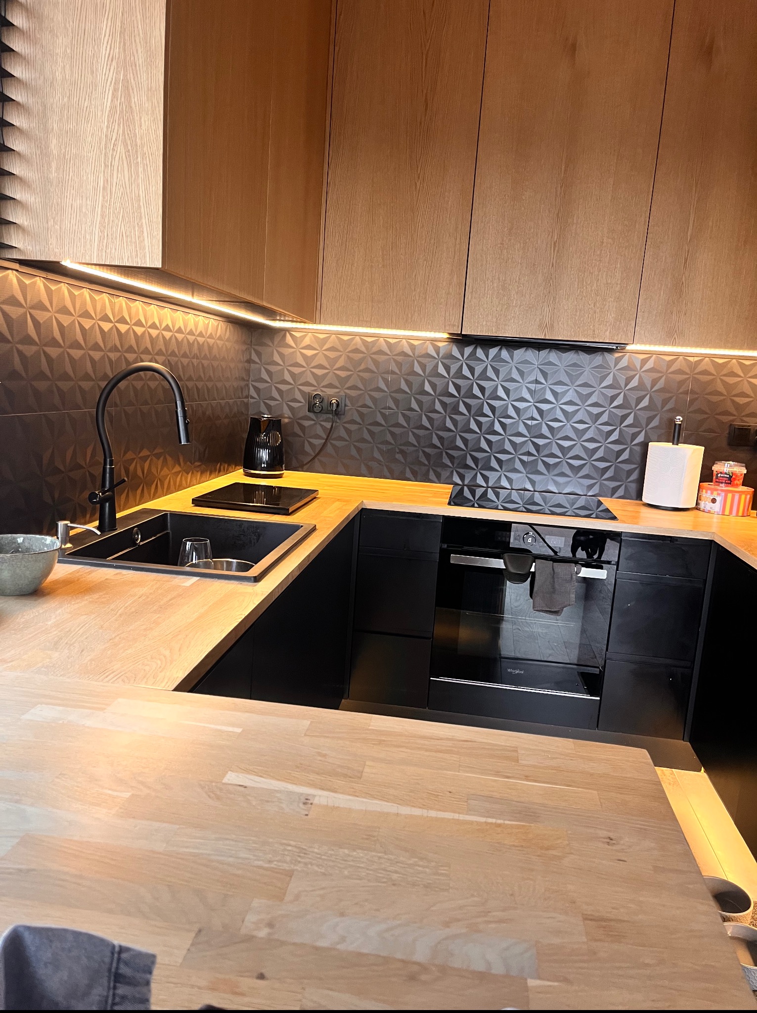 Nowoczesna kuchnia z drewnianymi szafkami, czarnym zlewem i geometryczną ścianą. Oświetlenie LED podkreśla unikalny design.