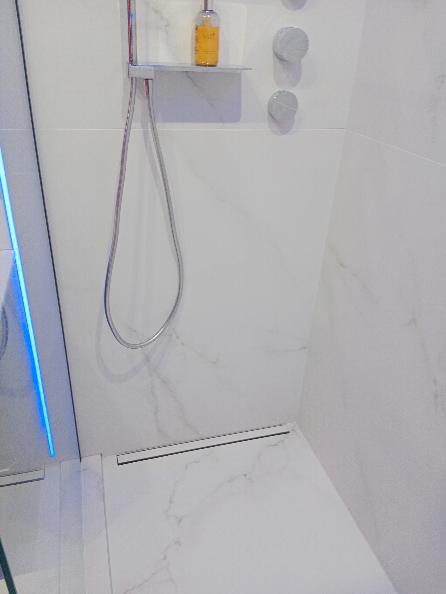 Kabina prysznicowa z białymi marmurkowymi płytkami, chromowaną armaturą i odpływem liniowym. Widoczna butelka kosmetyku na półce i podświetlane szkło.