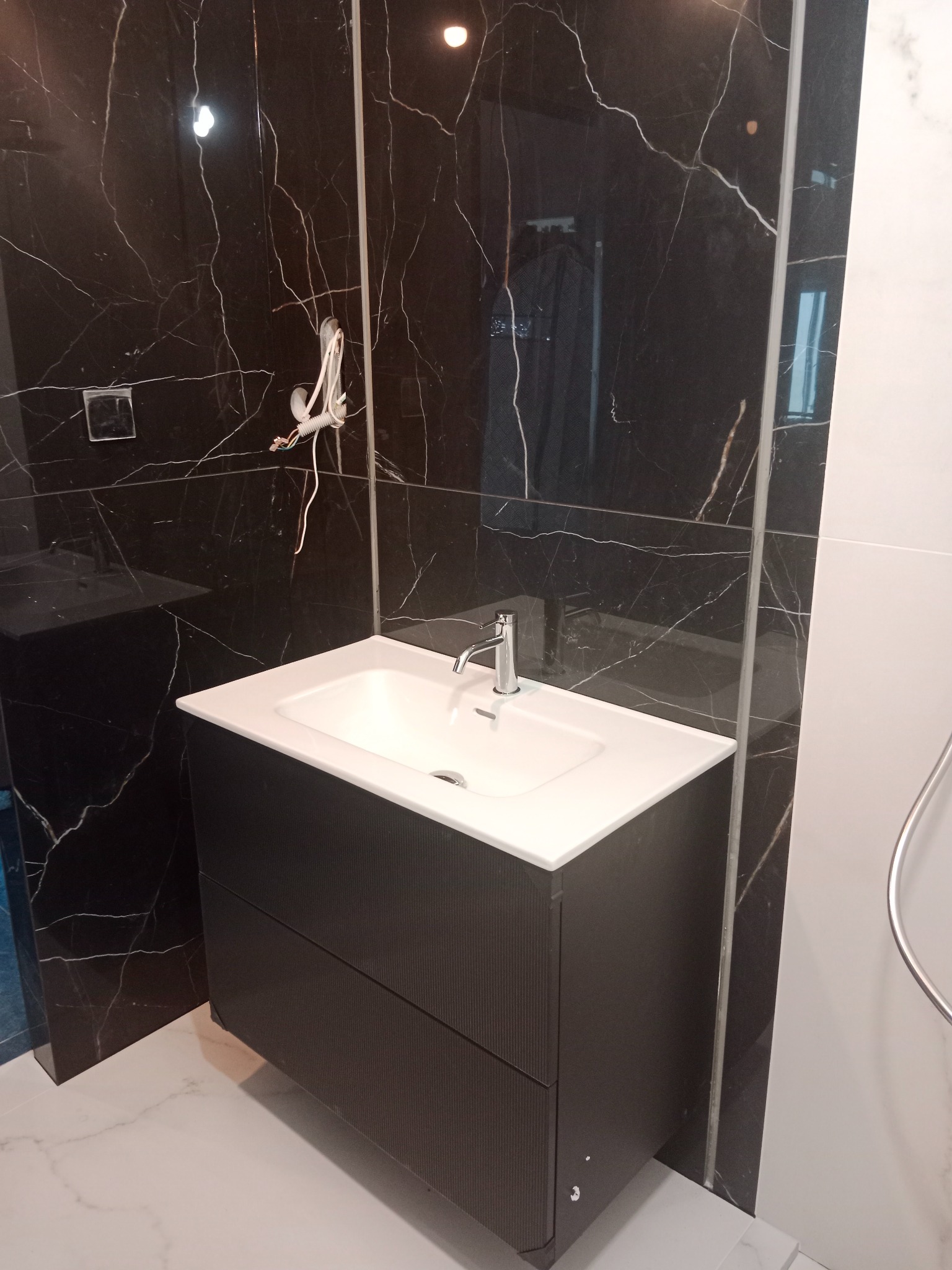Nowoczesna łazienka po remoncie: czarne marmurowe płytki, biała umywalka z szafką i armaturą. Widoczne przewody elektryczne przed ostatecznym wykończeniem.