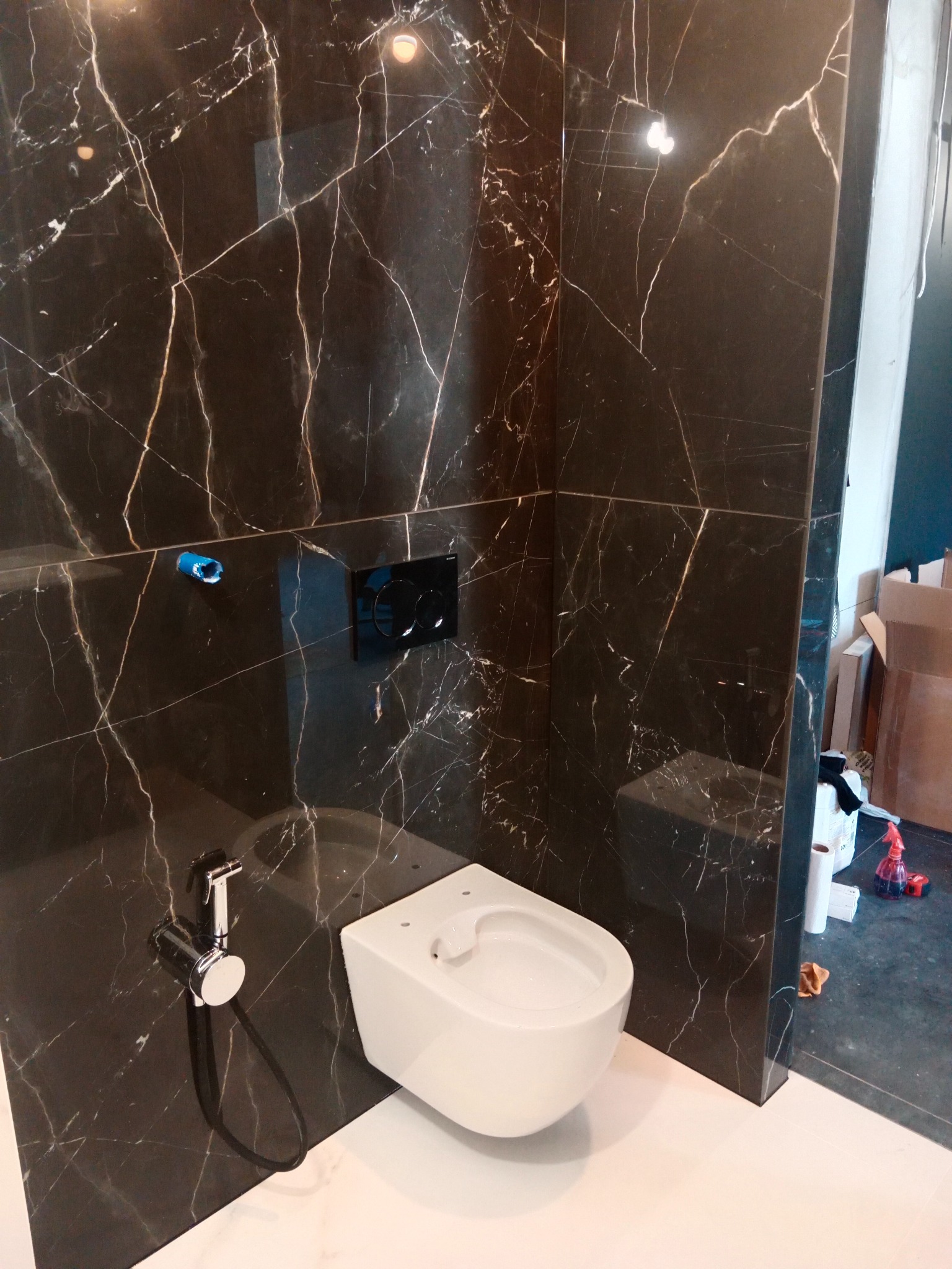 Nowoczesna toaleta podwieszana z bidetką, obudowana płytami imitującymi czarny marmur z białym użyleniem. Widoczne elementy instalacji i przycisk spłuczki.