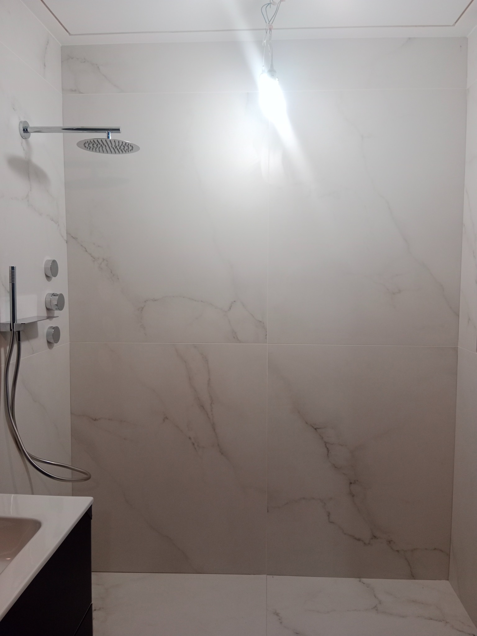 Nowoczesna kabina prysznicowa z marmurowymi płytkami, deszczownicą i baterią podtynkową. Minimalistyczny design, jasne oświetlenie, widoczny fragment wanny.