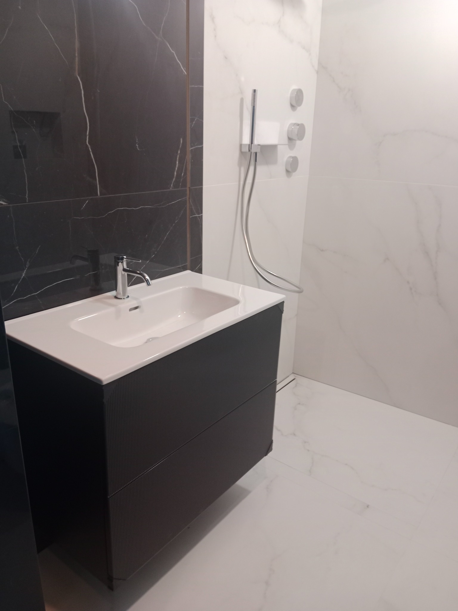 Nowoczesna łazienka: biała umywalka z chromowaną baterią na tle marmurowej ściany i kabiny prysznicowej z deszczownicą. Minimalistyczny design, eleganckie wykończenie.