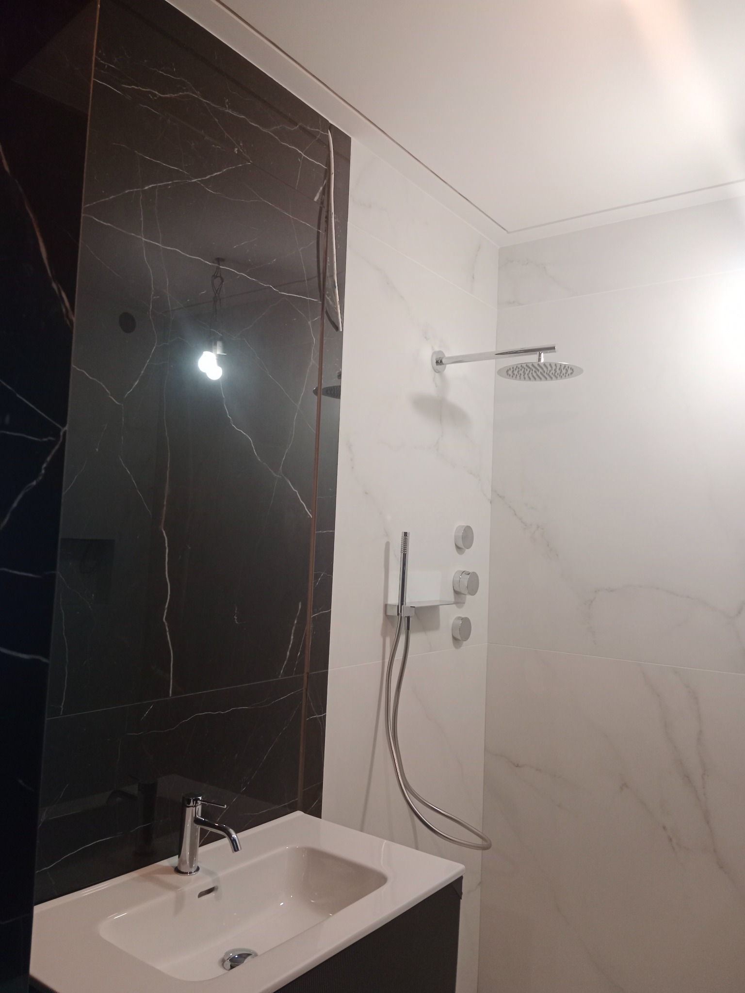 Nowoczesna łazienka z czarną marmurową ścianą i białą umywalką z baterią. Widoczny panel prysznicowy i deszczownica. Minimalistyczny design, jasne oświetlenie.