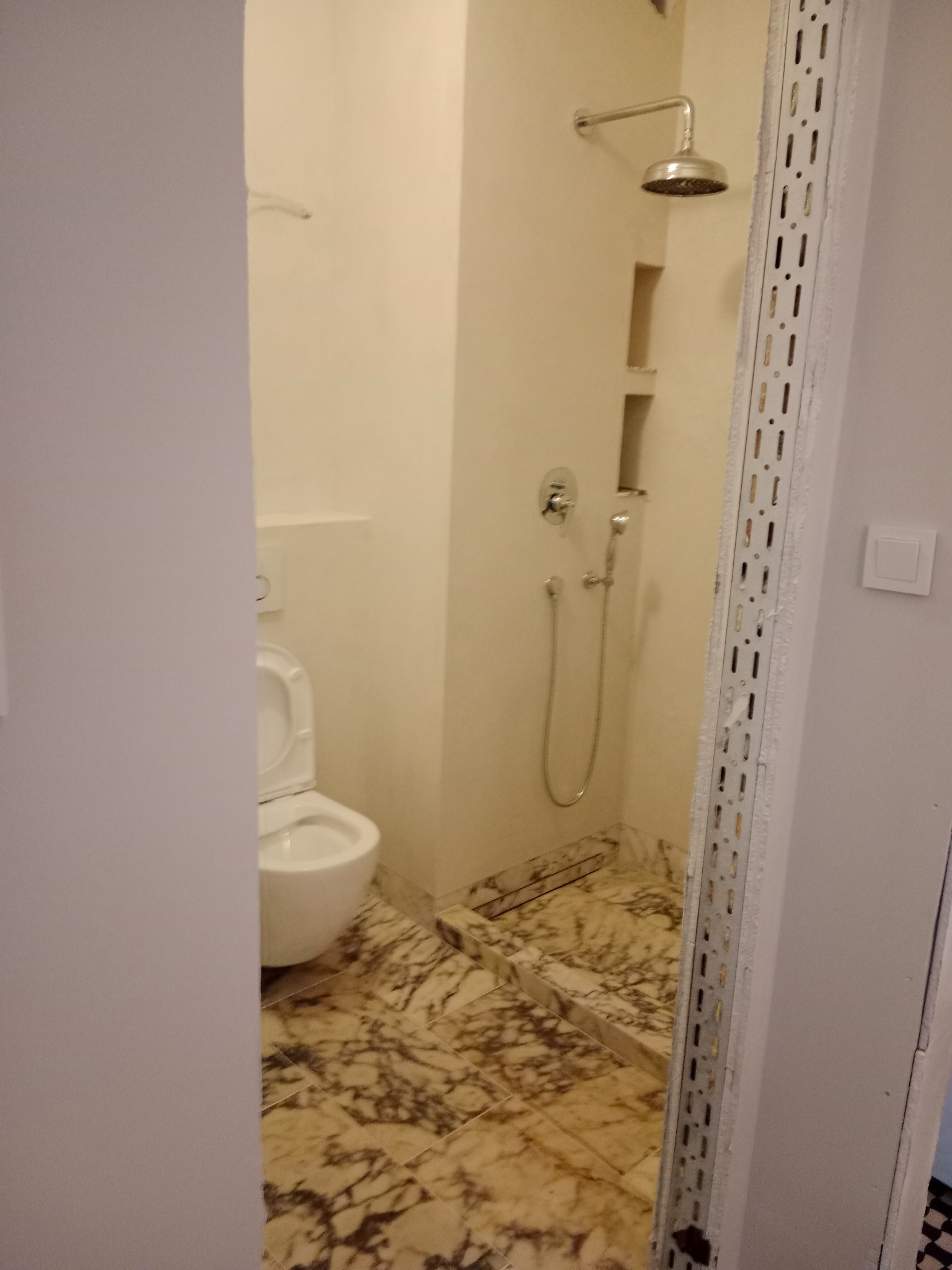 Wykończona łazienka z marmurową podłogą, widok zza framugi drzwi. Kabina prysznicowa z deszczownicą i toaleta. Widoczne półki w ścianie.