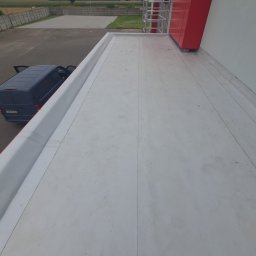 DKRM MAREK PIWOWARCZYK SPÓŁKA KOMANDYTOWO-AKCYJNA - Wykończony dach płaski z szarą membraną PVC, obróbki blacharskie w kolorze białym, zaparkowany samochód dostawczy z otwartymi drzwiami w tle.