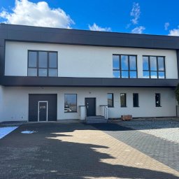 DKRM MAREK PIWOWARCZYK SPÓŁKA KOMANDYTOWO-AKCYJNA - Nowoczesny budynek biurowy z czarno-białą elewacją i dużymi oknami, widok z zewnątrz, kostka brukowa przed budynkiem, błękitne niebo.