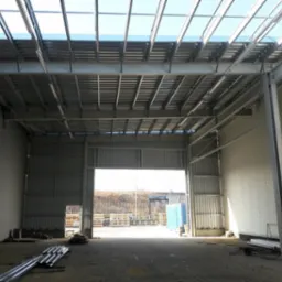 Wnętrze hali w trakcie budowy z widoczną konstrukcją stalową dachu i ścianami z płyt warstwowych. W tle widoczne otwarte wrota hali i teren zewnętrzny.