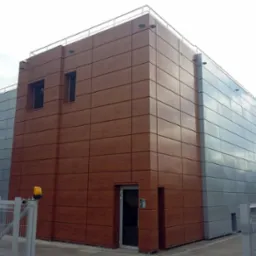 Nowoczesny budynek biurowy z elewacją z paneli w odcieniach brązu i szarości, widoczny z zewnątrz, z bramą wjazdową i elementami krajobrazu.