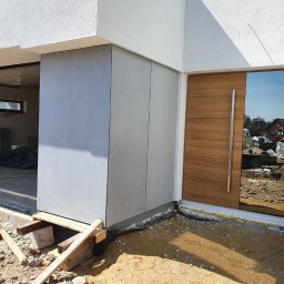 DKRM MAREK PIWOWARCZYK SPÓŁKA KOMANDYTOWO-AKCYJNA - Nowoczesna elewacja z szarych paneli i drewniane drzwi wejściowe z lustrzanym panelem, widok z zewnątrz na plac budowy, słoneczny dzień.