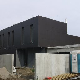 DKRM MAREK PIWOWARCZYK SPÓŁKA KOMANDYTOWO-AKCYJNA - Nowoczesny, dwukondygnacyjny dom z ciemną elewacją panelową i wysokimi oknami, otoczony betonowym ogrodzeniem w trakcie budowy.