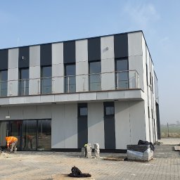 DKRM MAREK PIWOWARCZYK SPÓŁKA KOMANDYTOWO-AKCYJNA - Nowoczesny budynek z elewacją w pasy i balkonem. Na pierwszym planie pracownicy układają kostkę brukową. Widoczny podnośnik koszowy i materiały budowlane.