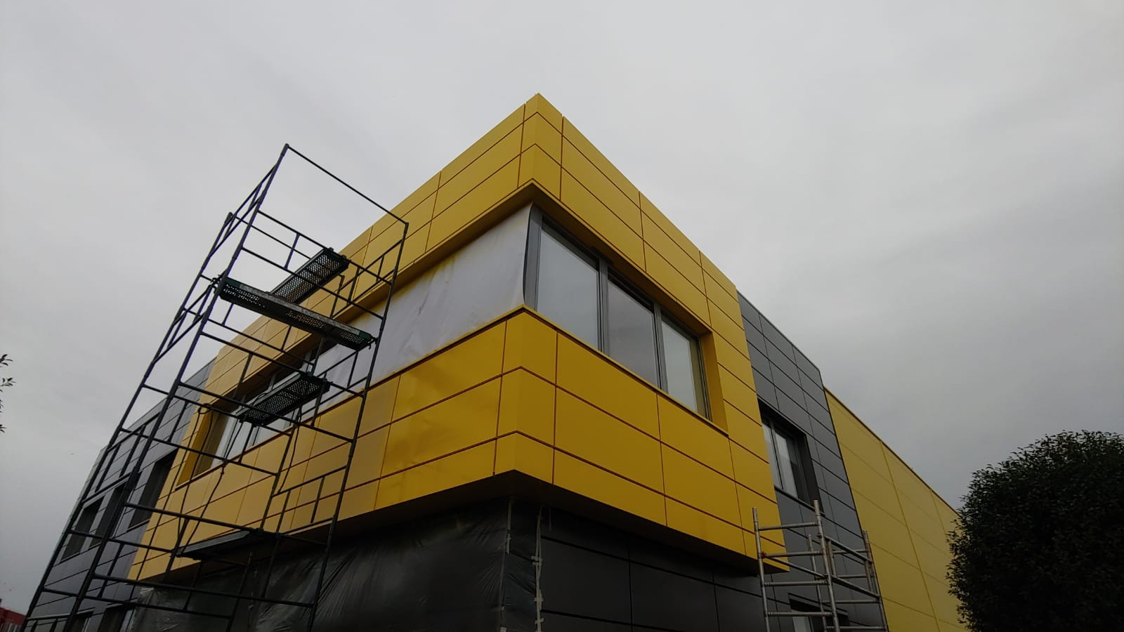 Żółto-szara elewacja budynku z panelami, widoczne rusztowania. Ujęcie z dołu, pochmurne niebo w tle. Geometryczny design elewacji.