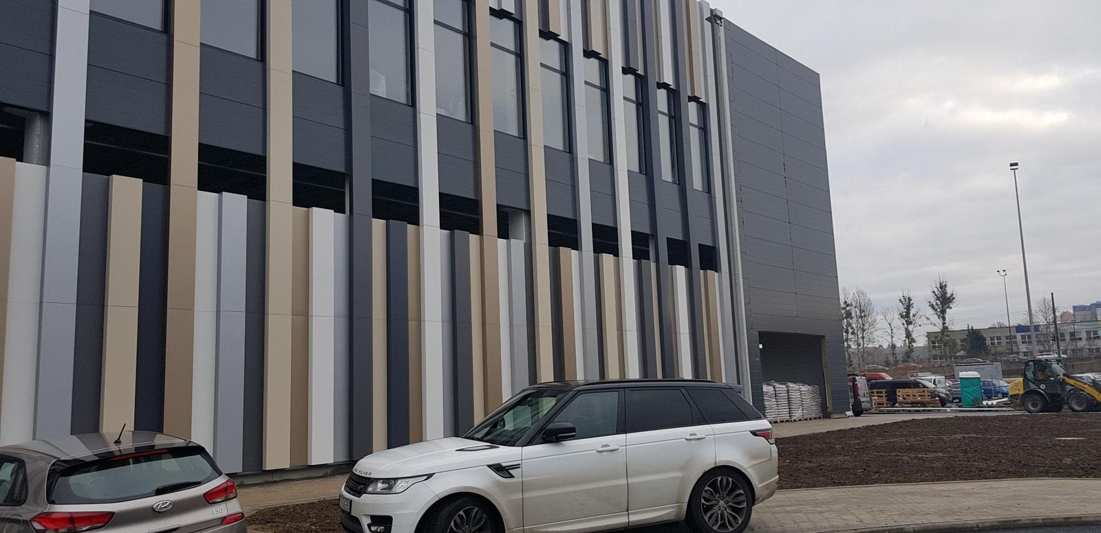 Nowoczesna elewacja hali z pionowymi panelami w odcieniach szarości i beżu, z zaparkowanymi samochodami osobowymi na pierwszym planie. Widoczne okna i fragment placu budowy.