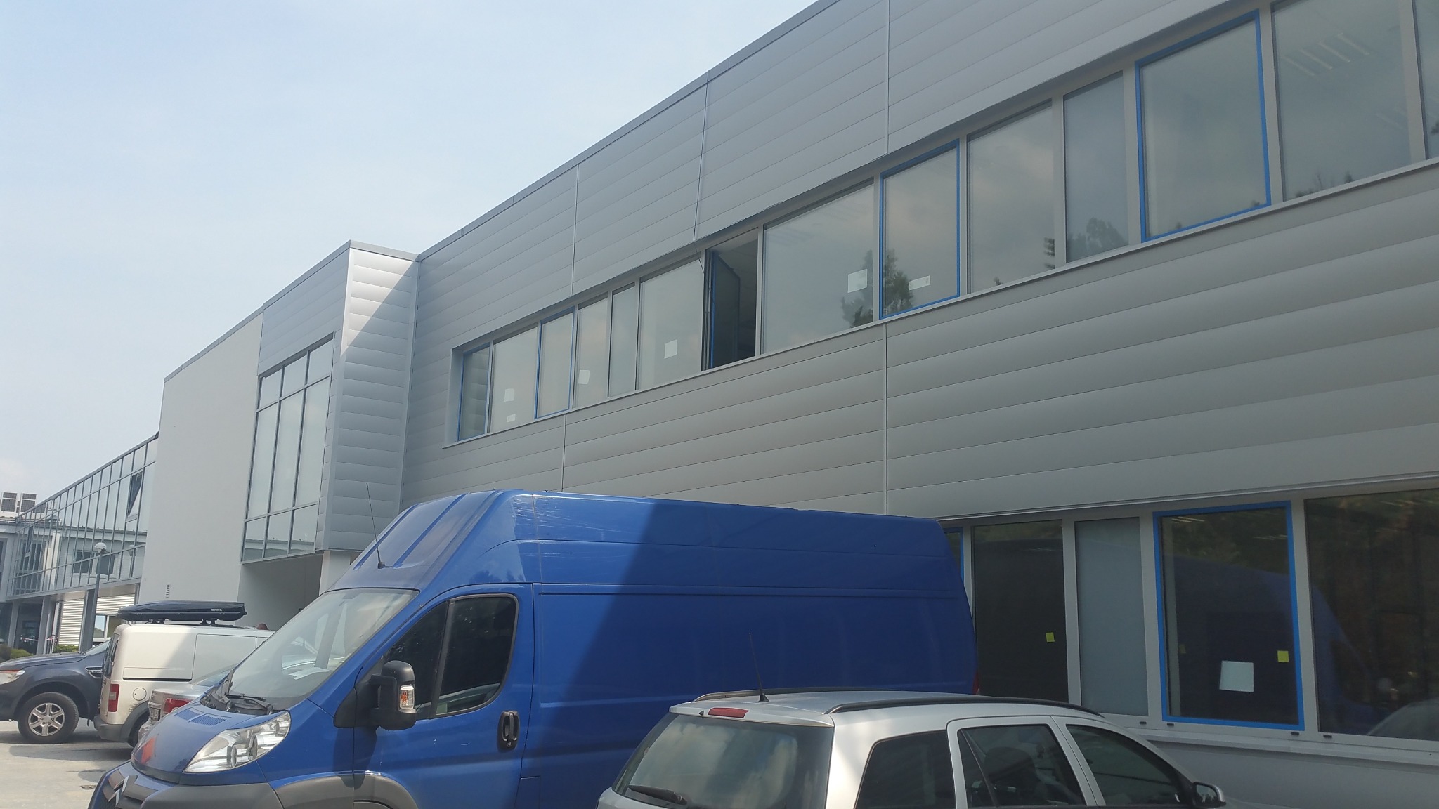 Budynek hali z szarej blachy trapezowej z oknami, częściowo zasłonięty przez niebieski samochód dostawczy i srebrny samochód osobowy. Widoczne inne zaparkowane pojazdy.