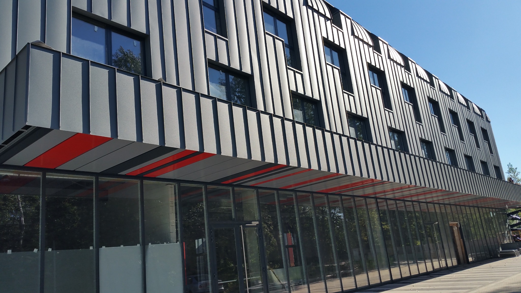 Nowoczesna hala z szarej blachy trapezowej, z rzędem okien i przeszklonym parterem z czerwonym akcentem. Widok z dołu, błękitne niebo w tle.