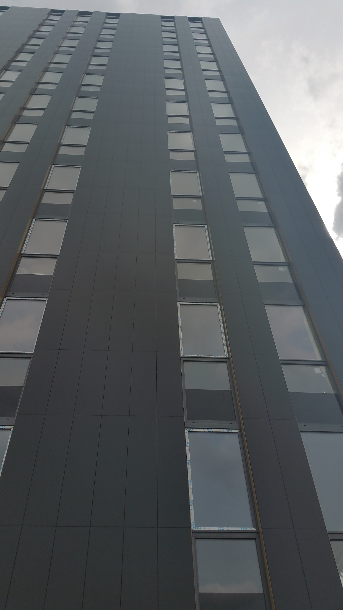 Widok z dołu na szary, wysoki budynek z rzędami okien. Elewacja wykonana z paneli. Pochmurne niebo w tle. Ujęcie z perspektywy żabiej.