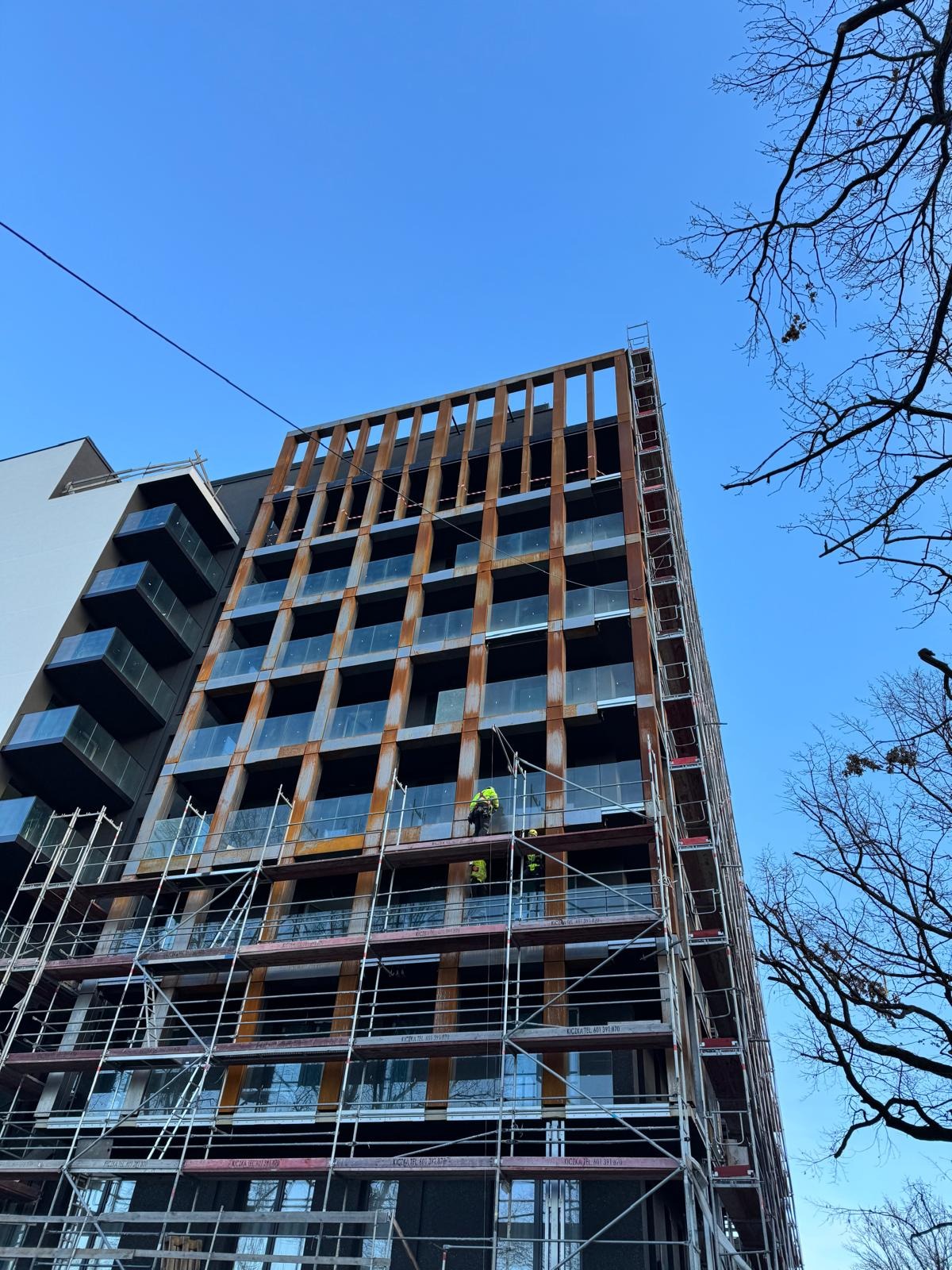 Wielopiętrowy budynek w trakcie prac elewacyjnych, z rusztowaniami i pracownikami w żółtych kamizelkach, na tle błękitnego nieba i drzew bez liści.