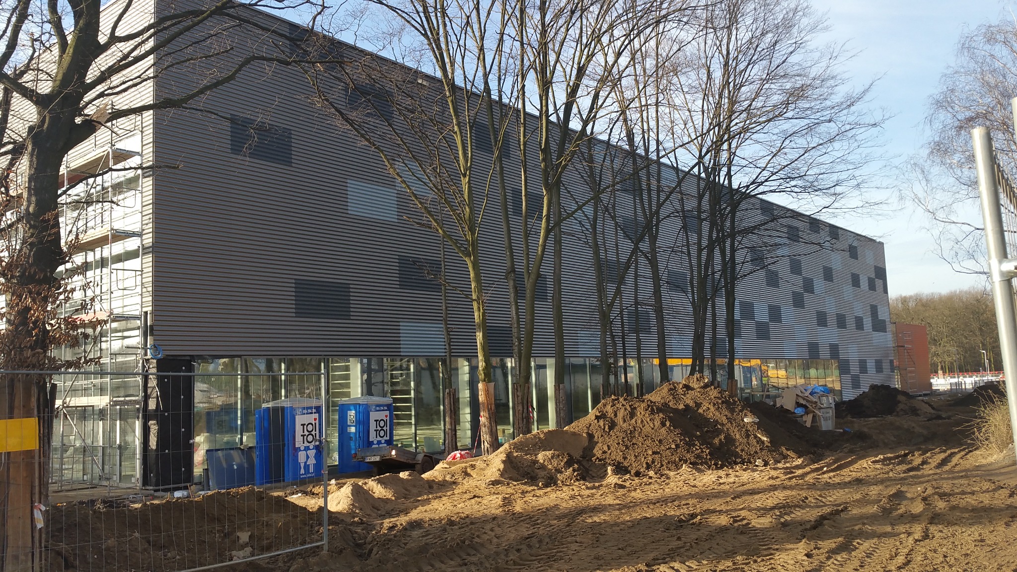 Nowoczesna hala w trakcie budowy z szarą elewacją z blachy trapezowej, widoczne rusztowania i teren budowy, drzewa bez liści w pierwszym planie.