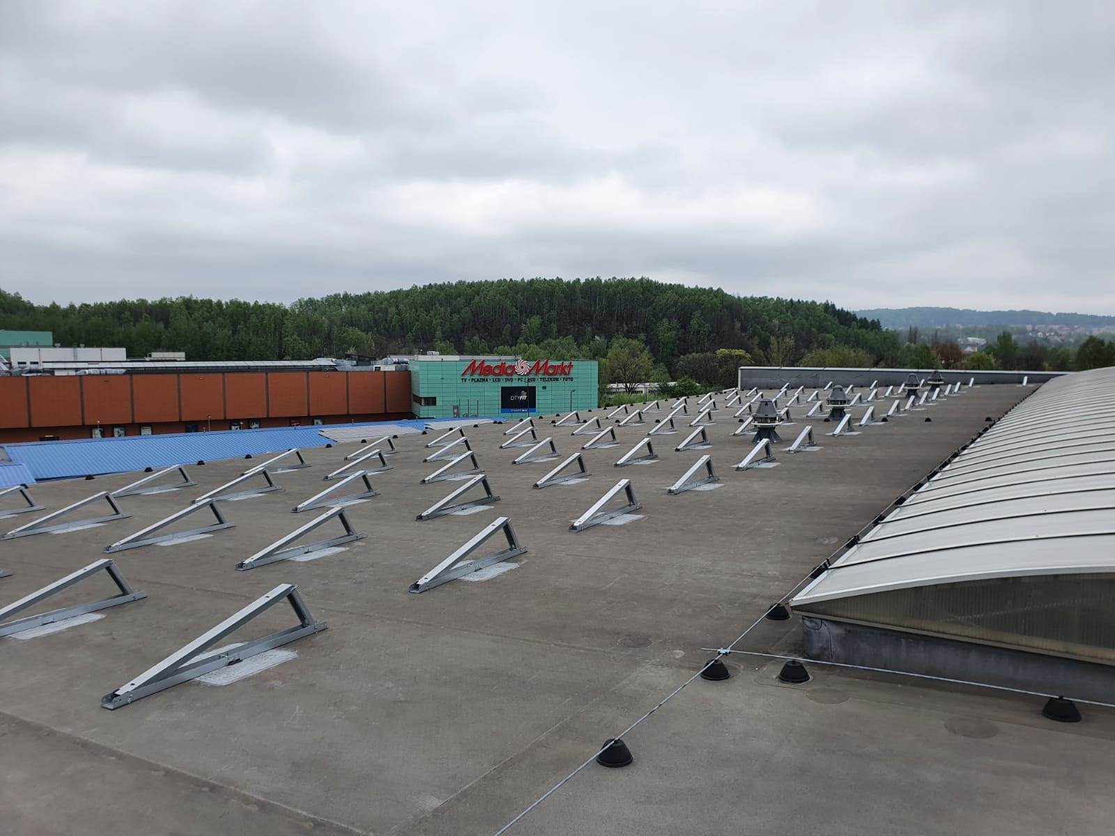 Montaż konstrukcji wsporczej pod panele fotowoltaiczne na dachu budynku handlowego Media Markt, widok z góry, pochmurne niebo.
