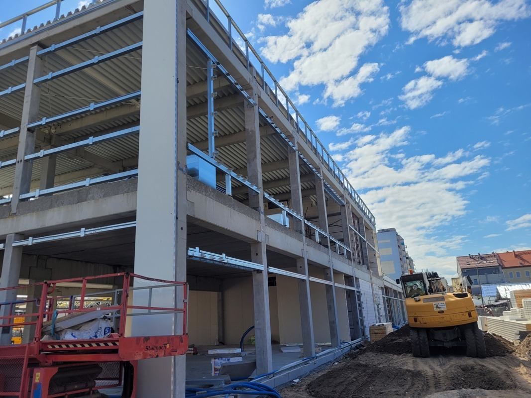 Konstrukcja hali w budowie, widoczne stalowe elementy i betonowe słupy, na zewnątrz sprzęt budowlany i materiały izolacyjne. Słoneczny dzień, błękitne niebo.