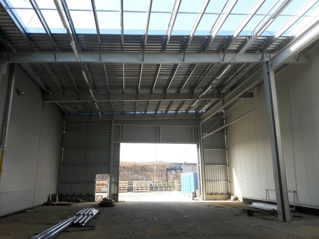 Wnętrze hali w trakcie budowy z widoczną konstrukcją stalową dachu i ścianami z płyt warstwowych. W tle widoczne otwarte wrota hali i teren zewnętrzny.