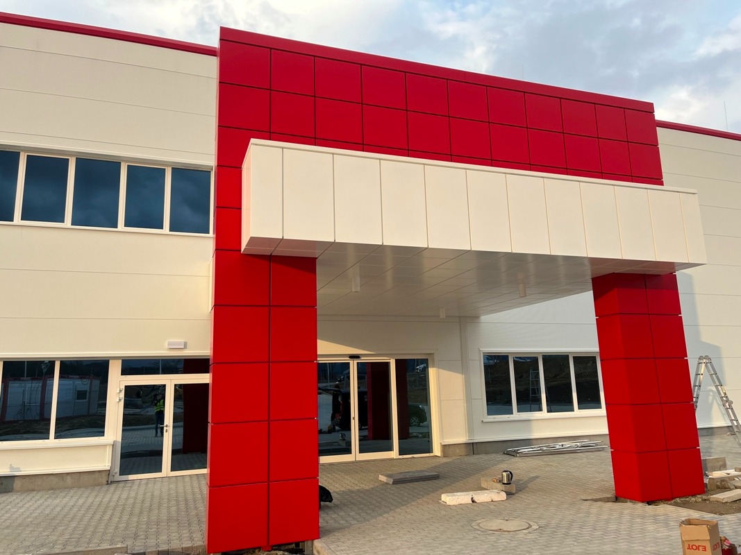 Nowoczesna hala z elewacją z płyt warstwowych w kolorach białym i czerwonym. Widoczne okna, drzwi wejściowe i częściowo ułożona kostka brukowa przed budynkiem.
