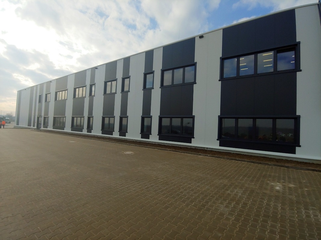 Nowoczesna hala przemysłowa z elewacją w pasy, czarno-biała, z rzędem okien. Widok z zewnątrz, na pierwszym planie bruk. Błękitne niebo z chmurami w tle.