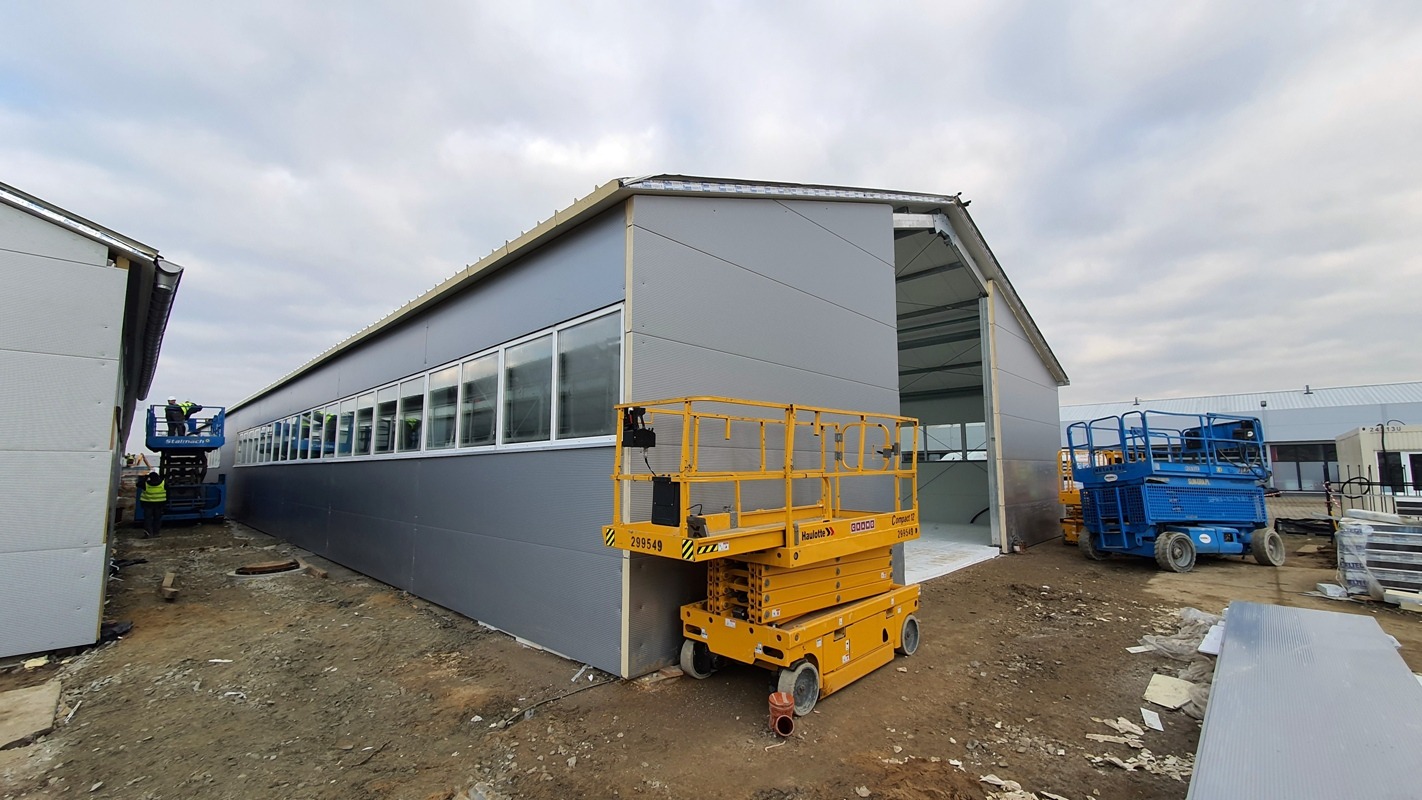 Hala w trakcie budowy z szarymi ścianami z płyt warstwowych, żółty podnośnik nożycowy na pierwszym planie, niebieski podnośnik w tle, pochmurne niebo.