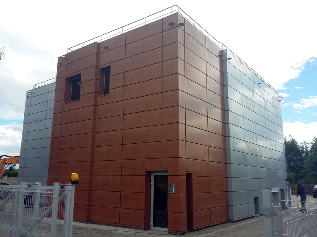 Nowoczesny budynek biurowy z elewacją z paneli w odcieniach brązu i szarości, widoczny z zewnątrz, z bramą wjazdową i elementami krajobrazu.