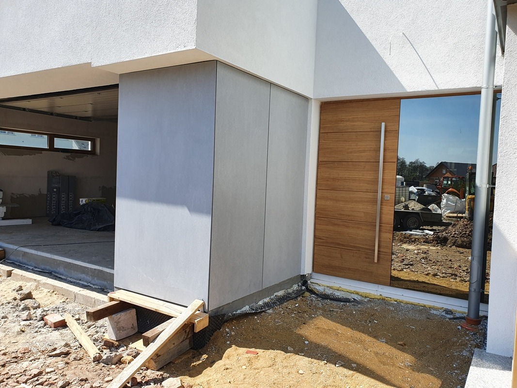 Nowoczesna elewacja z szarych paneli i drewniane drzwi wejściowe z lustrzanym panelem, widok z zewnątrz na plac budowy, słoneczny dzień.