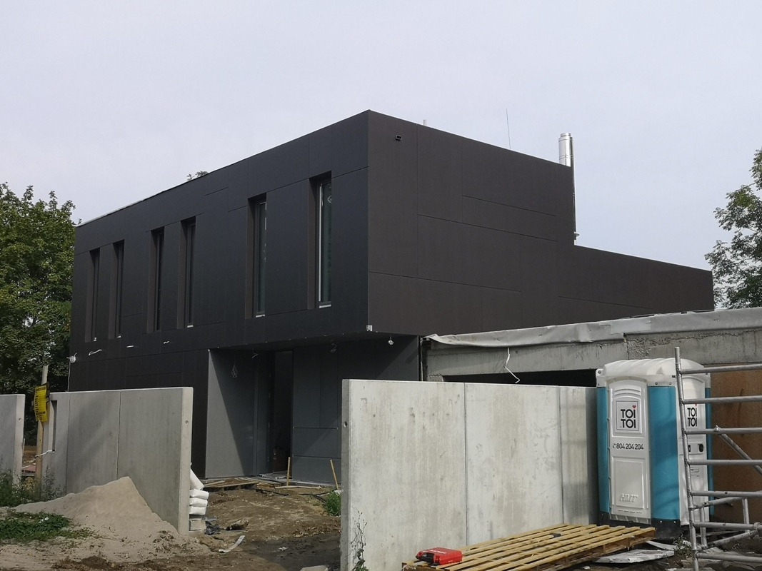 Nowoczesny, dwukondygnacyjny dom z ciemną elewacją panelową i wysokimi oknami, otoczony betonowym ogrodzeniem w trakcie budowy.
