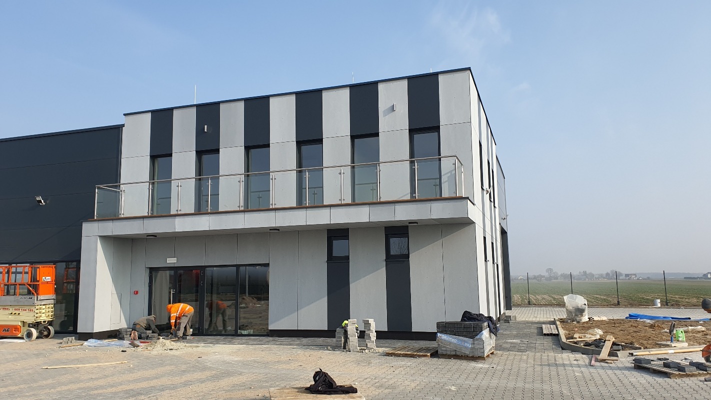 Nowoczesny budynek z elewacją w pasy i balkonem. Na pierwszym planie pracownicy układają kostkę brukową. Widoczny podnośnik koszowy i materiały budowlane.