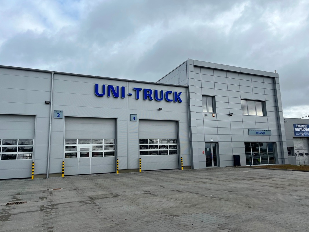 Szara hala UNI-TRUCK z bramami nr 3 i 4, widoczne biuro recepcyjne, oświetlenie zewnętrzne i kostka brukowa na placu przed budynkiem. Pochmurne niebo w tle.