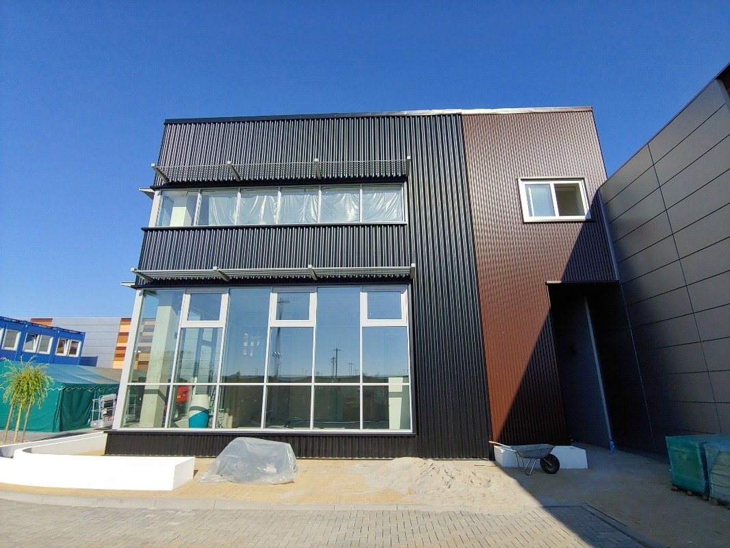 Nowoczesny budynek biurowy z elewacją z blachy trapezowej w kolorze grafitowym i brązowym, duże okna, błękitne niebo w tle. Widoczny wózek budowlany i materiały budowlane.