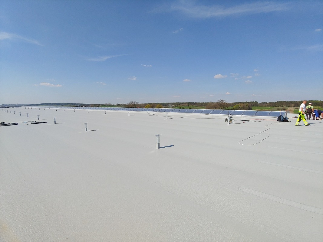 Biały dach hali z zamontowanymi panelami słonecznymi w oddali. Na dachu widoczni pracownicy w strojach roboczych i metalowe wsporniki.