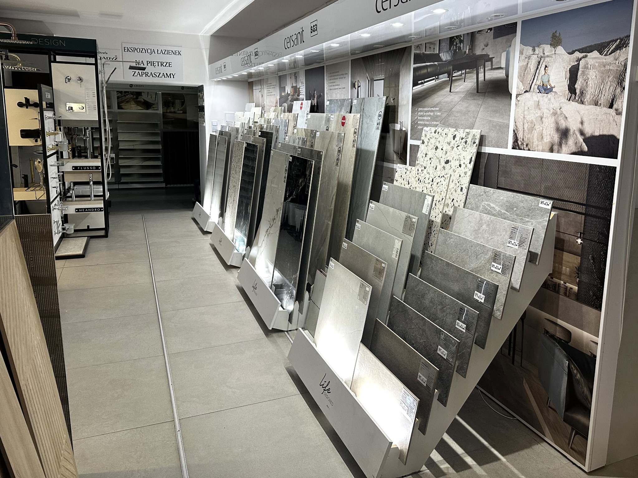 Ekspozycja płytek Cersanit w salonie łazienek, prezentująca różnorodność wzorów i faktur. Na ścianie grafika z aranżacją wnętrza łazienki.