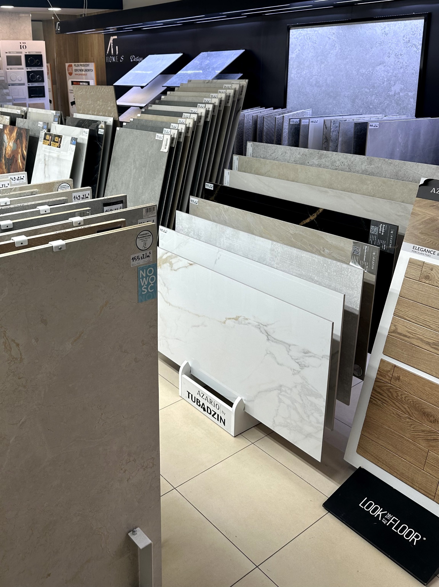 Ekspozycja płytek ceramicznych w sklepie: różnorodne wzory, faktury i kolory, od marmurkowych po imitujące drewno. Widoczne logo producenta Azario by Tubądzin.