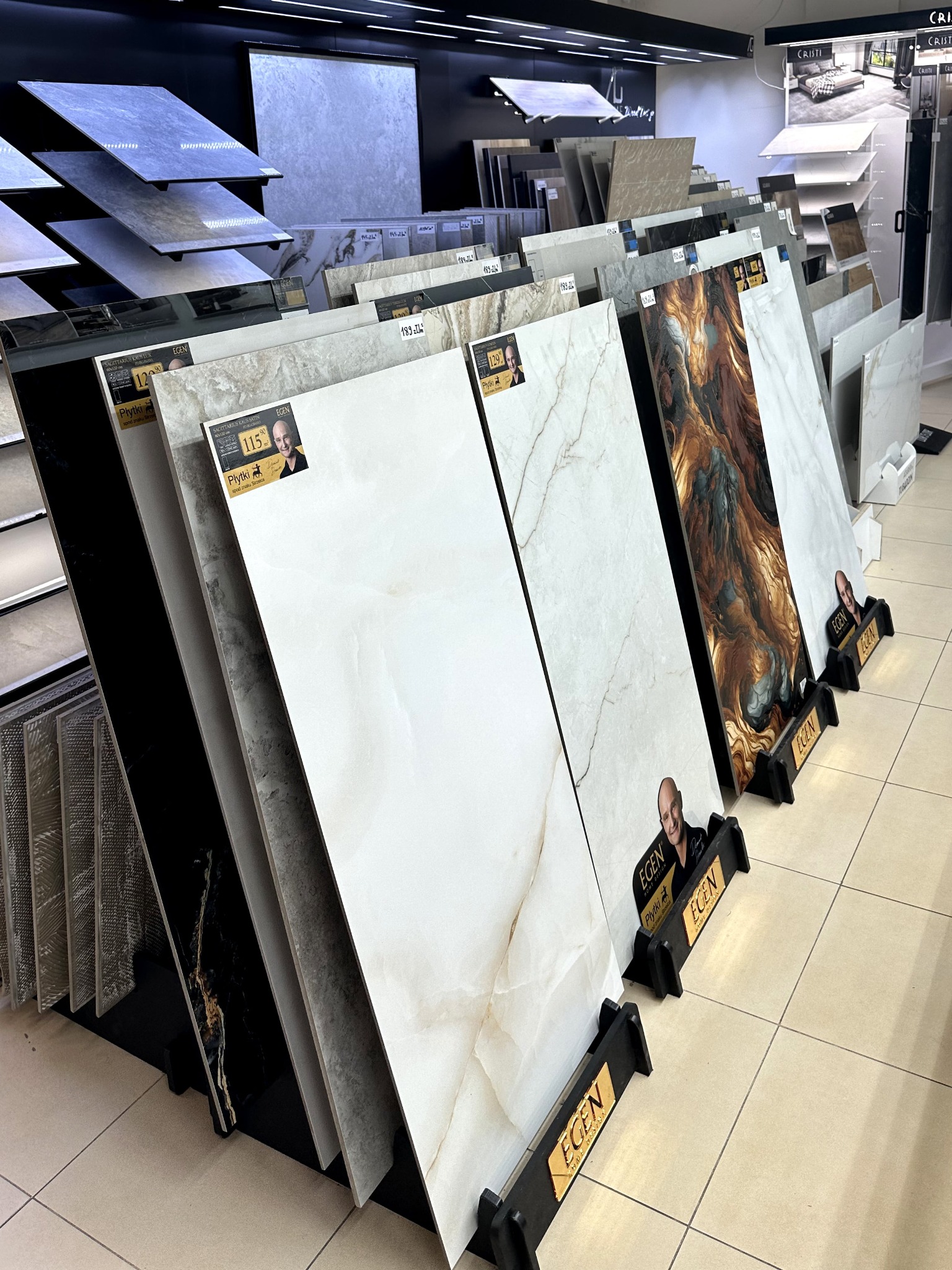 Ekspozycja różnorodnych płytek w salonie: imitacja marmuru, czarne z żyłami, w odcieniach beżu i szarości. Na każdym stojaku widoczna cena i logo producenta.
