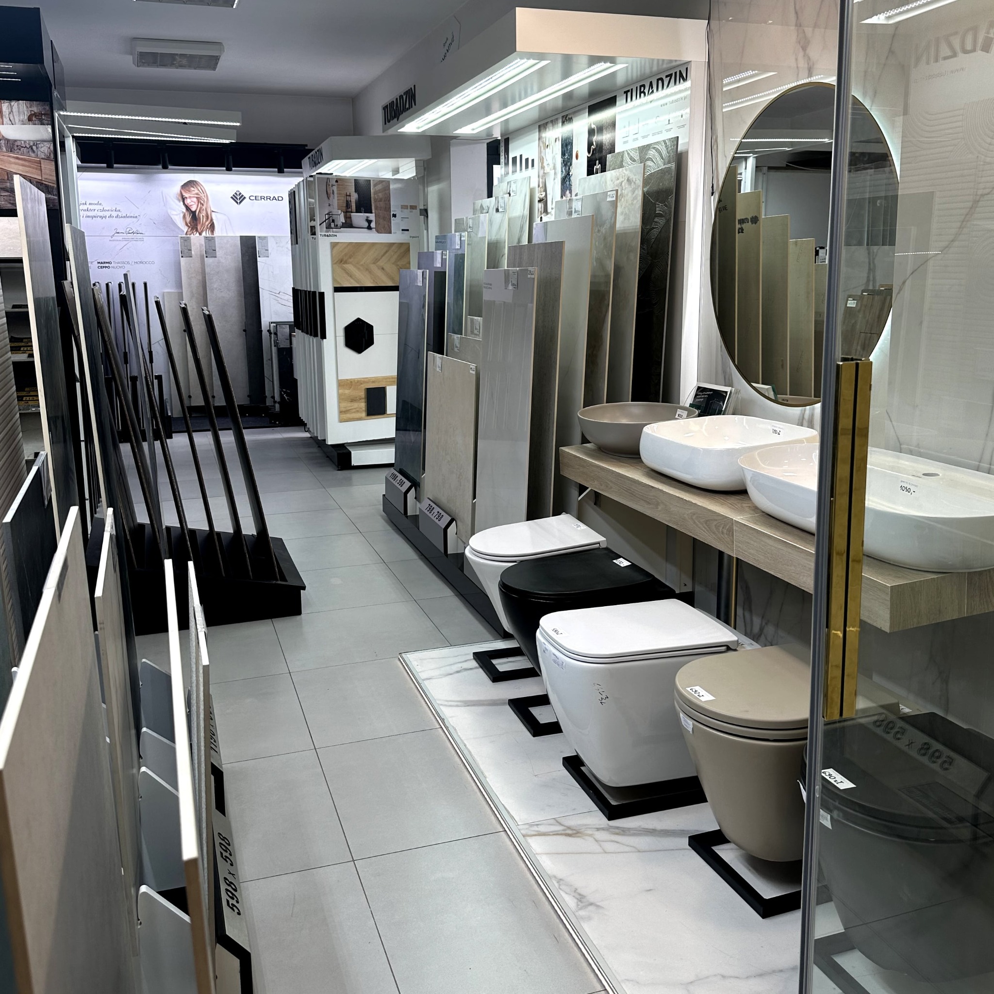 Ekspozycja płytek i armatury łazienkowej w salonie: umywalki nablatowe, toalety w różnych kolorach, duże formaty płytek. Widoczne logo Cerrad i Tubądzin.