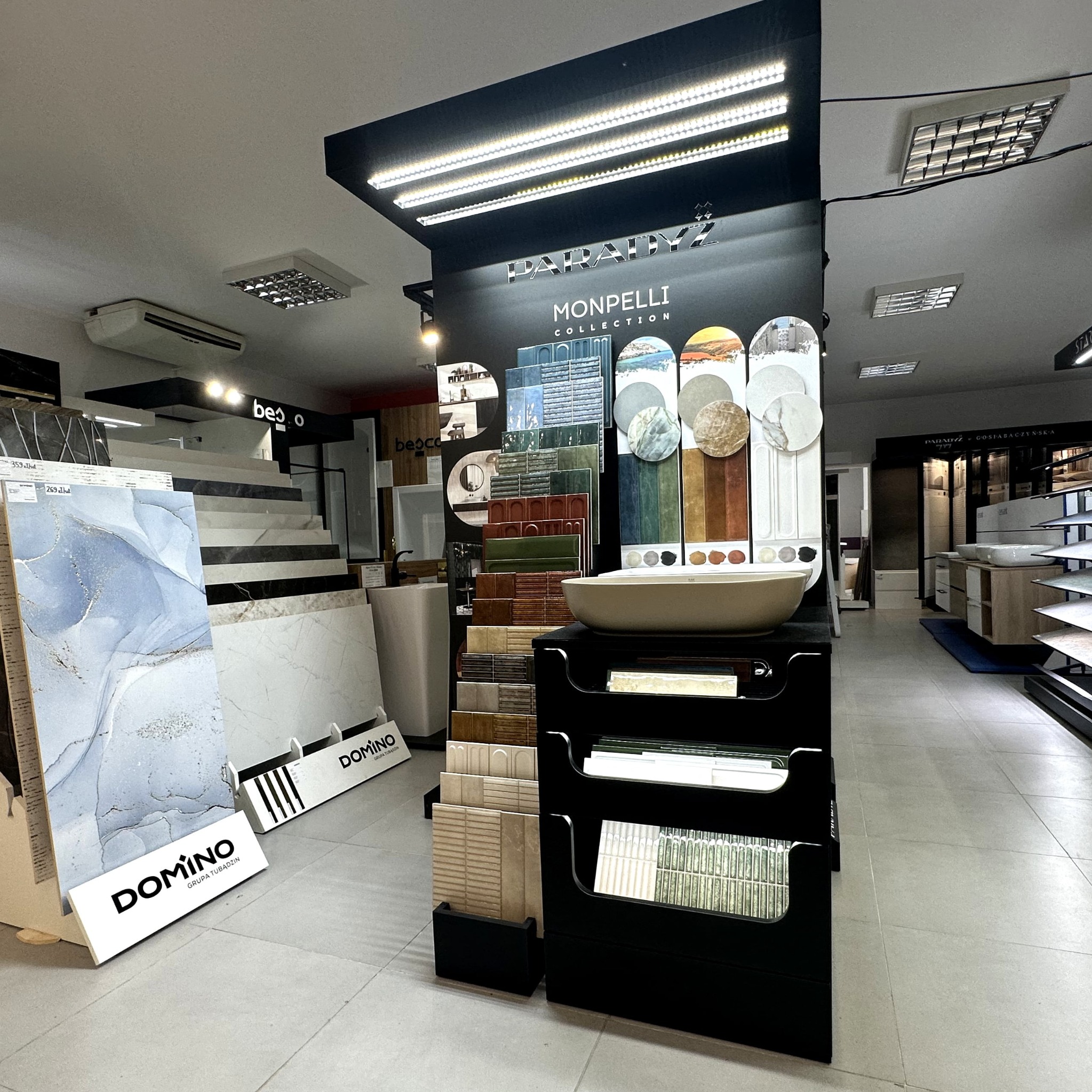 Ekspozycja płytek i umywalki z kolekcji Monpelli marki Paradyż w salonie, z widocznymi wzorami i kolorami. Płytki Domino. Oświetlenie sufitowe.