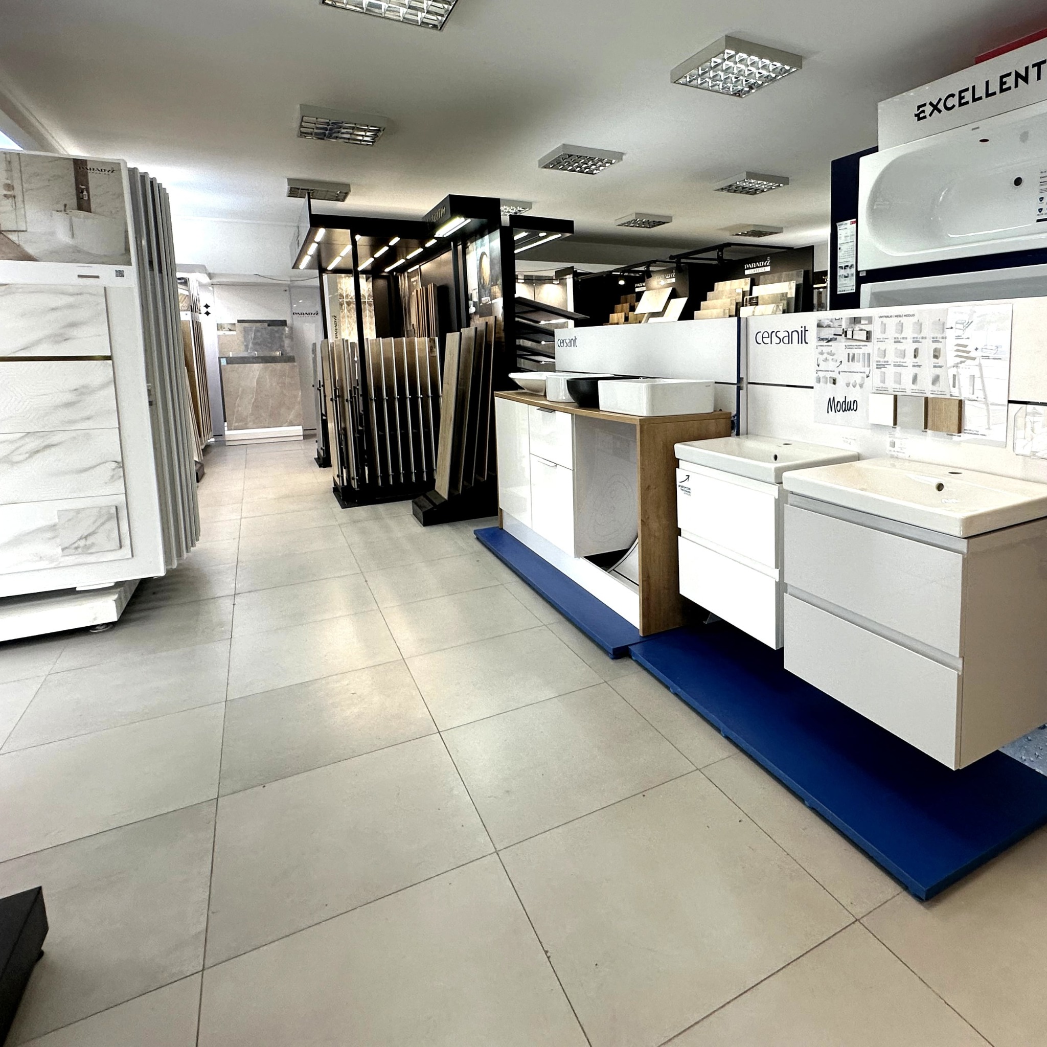 Ekspozycja w salonie łazienek: umywalki z szafkami Cersanit Moduo, płytki ceramiczne imitujące marmur i drewno. Oświetlenie sufitowe.