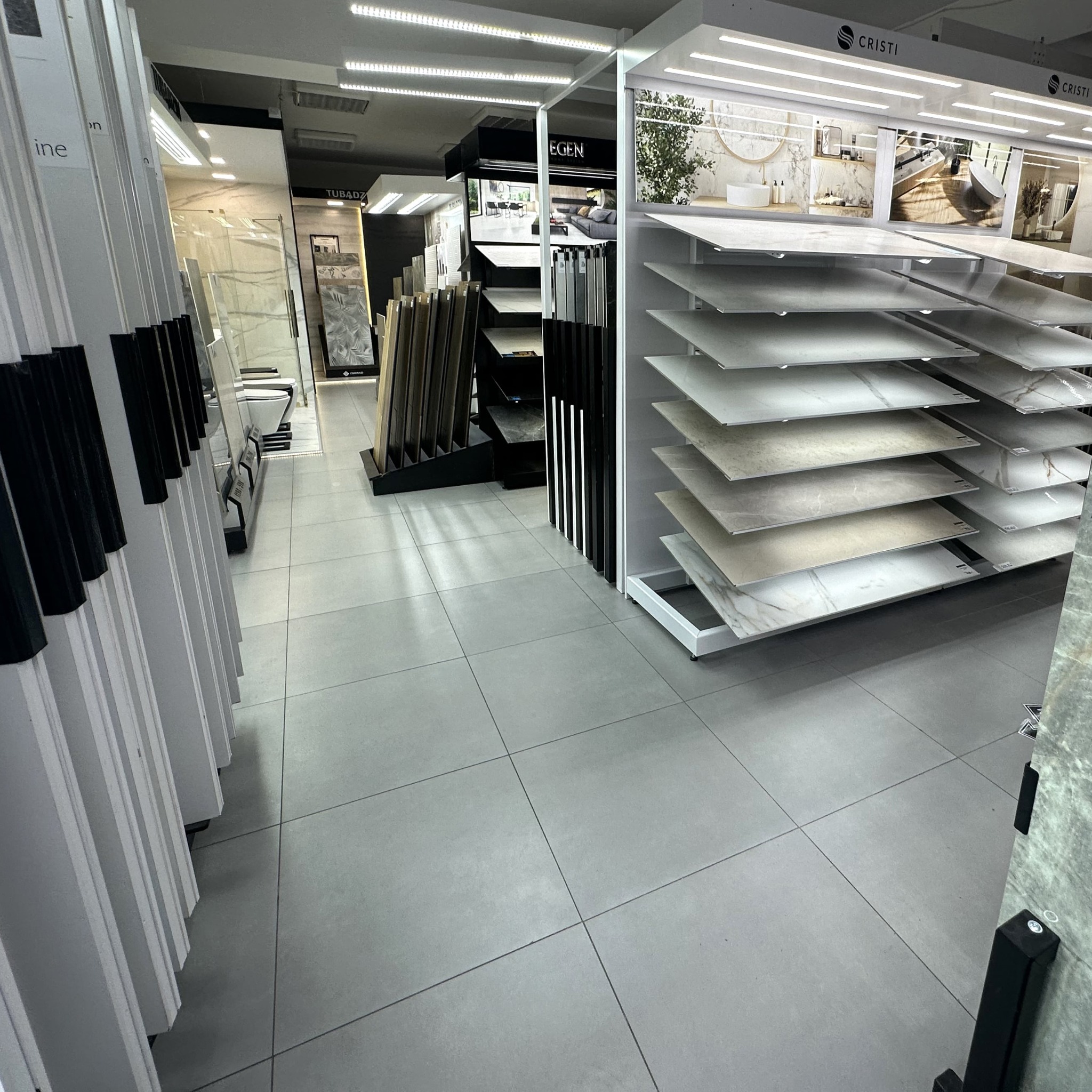 Ekspozycja płytek w salonie: różnorodne wzory i faktury na półkach, szare płytki podłogowe. Widoczne inne ekspozycje w tle, oświetlenie LED.