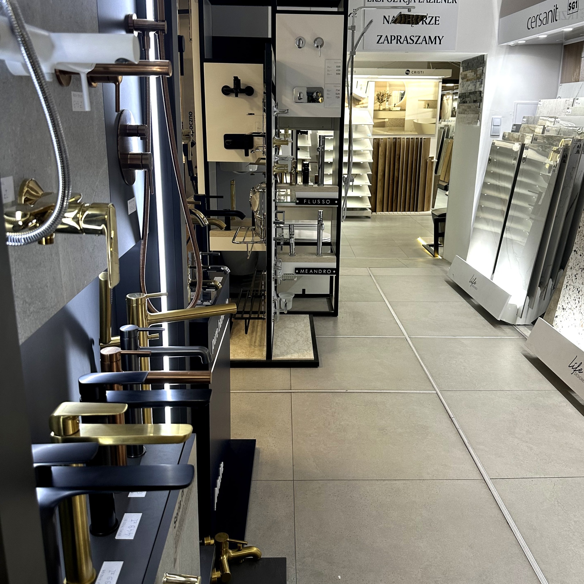 Ekspozycja baterii łazienkowych w salonie: złote, czarne i miedziane modele na tle płytek i armatury. Widoczny napis 'Zapraszamy' i logo Cersanit.